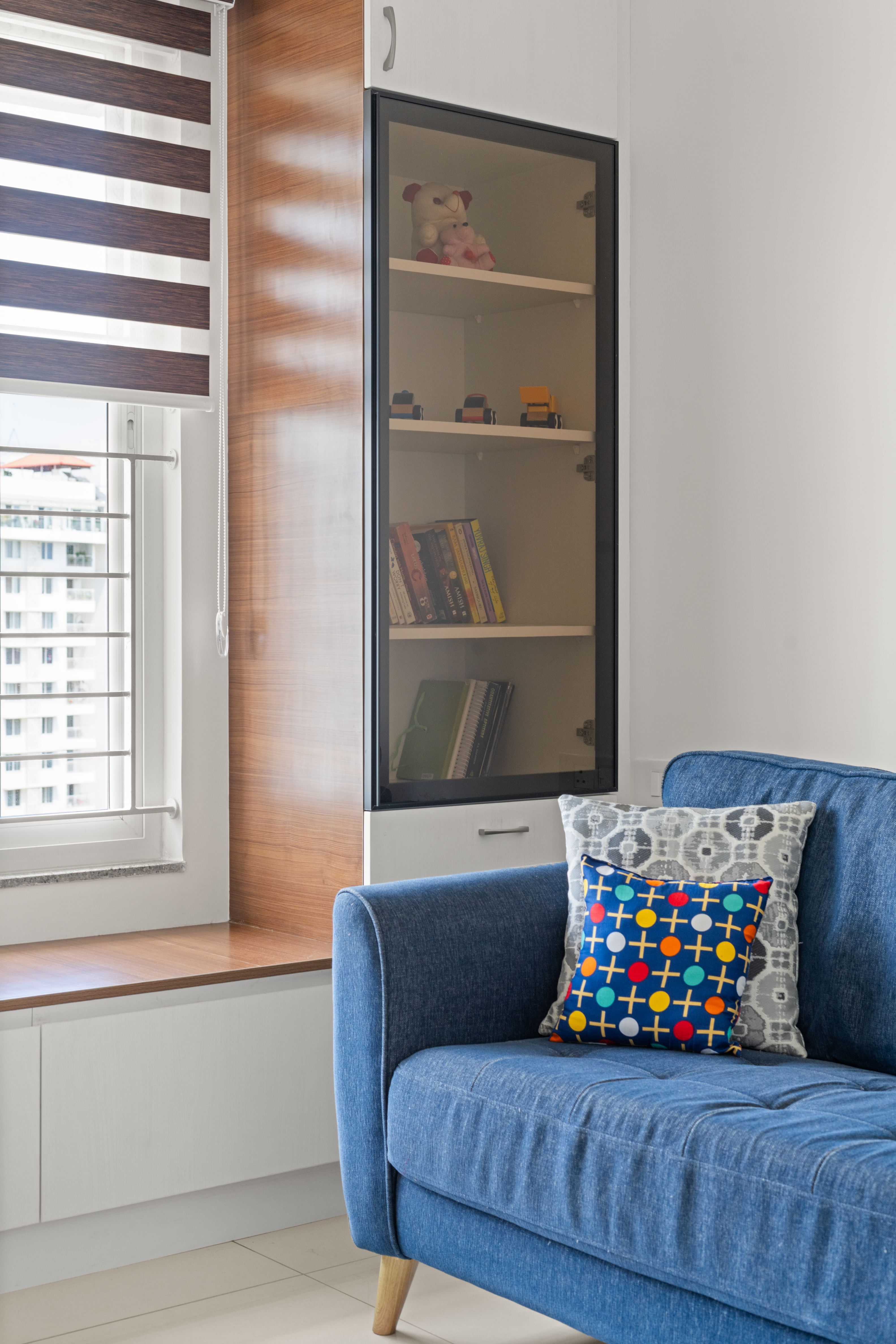 Modern living room with blue fabric sofa, walnut wood laminate window seat, and white laminate cabinetry of 2 BHK in Purva Palm Beach Hennur Bengaluru | Material Depot